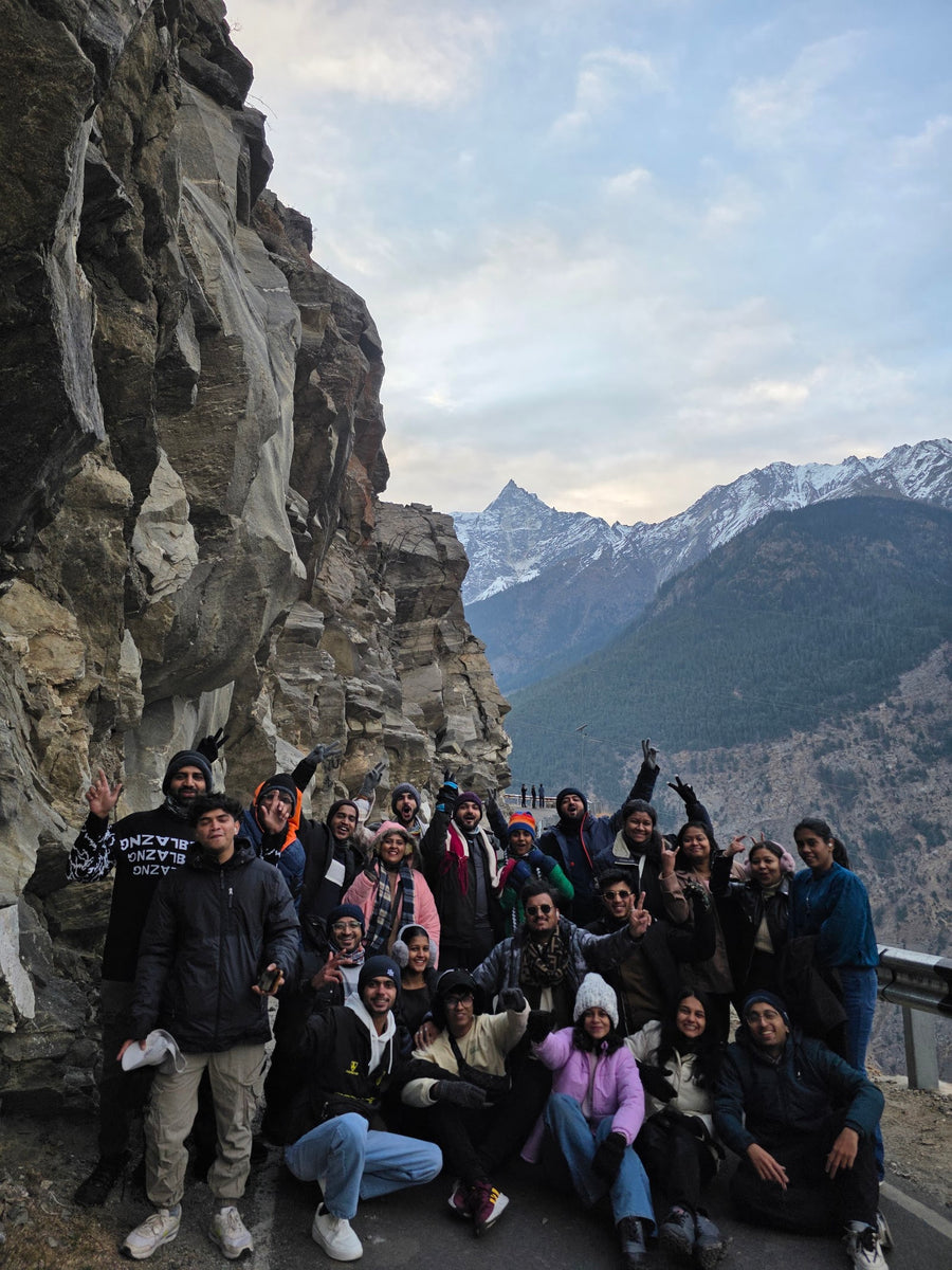 A large group in winter jackets on a cliff road with snow peaks behind