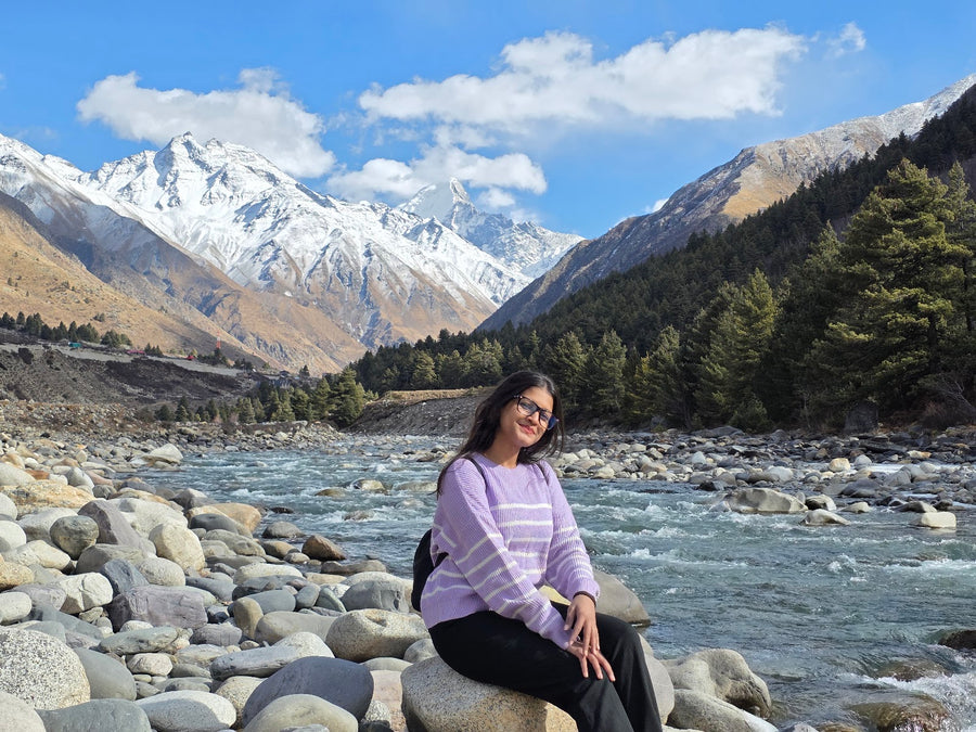 A traveller sitting on river boulders with pine forest and snow peaks behind
