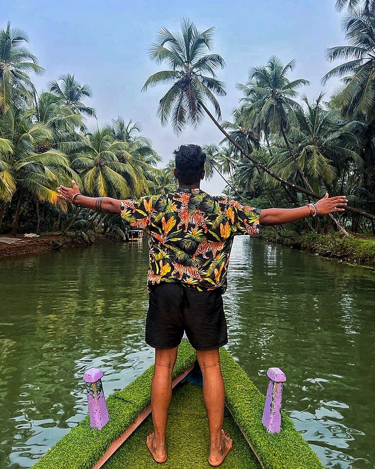 A traveller standing with arms wide on the bow of a boat among palms