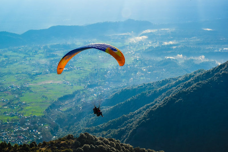 A paraglider with an orange and blue wing flying above the green Kangra valley near Bir Billing