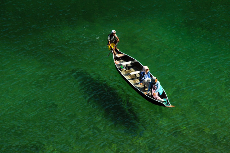 A wooden boat with three people on the clear green Umngot river at Dawki, its shadow visible on the riverbed