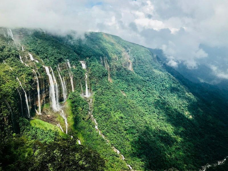 Streams of water running down a green cliff face into a cloud-filled valley in the Khasi Hills near Shillong